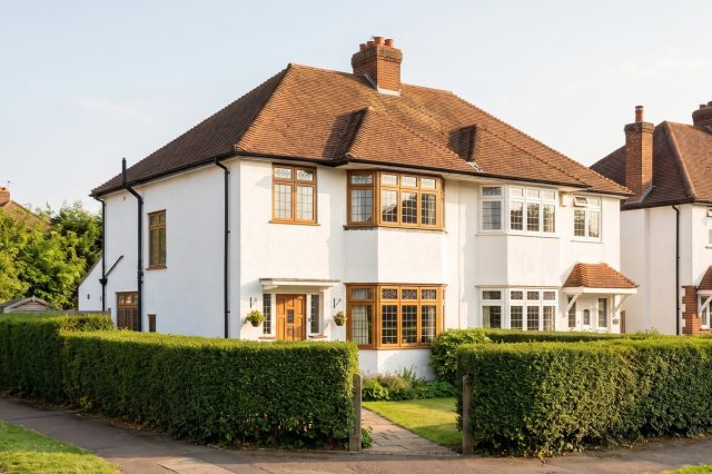 A 1930s British semi-detached house with white render, bay windows, a hipped clay tile roof and a neat privet hedge, in warm sunny afternoon light