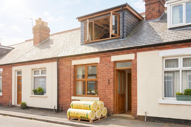 A row of modest British houses in bright sunshine with one loft window open, and a foreground of neatly rolled insulation resting by a doorway