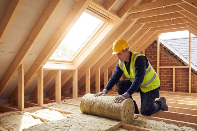 A bright British loft with a tradesman in a yellow hard hat and hi-vis vest unrolling fresh mineral wool insulation between joists