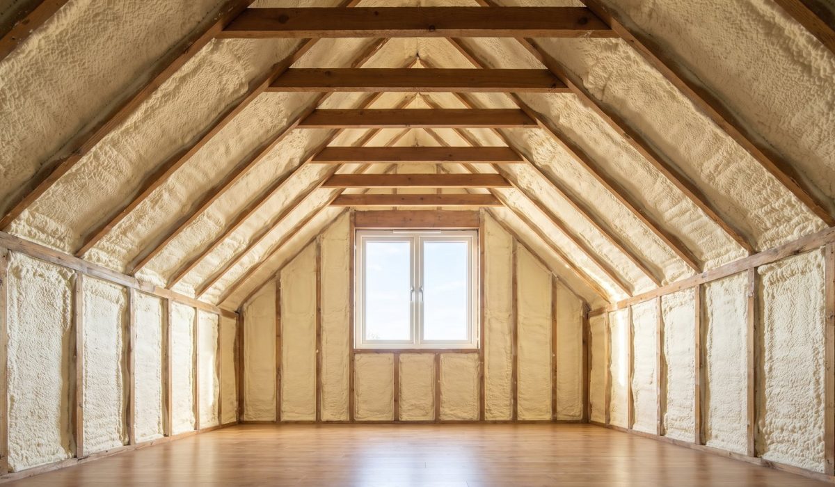 A British loft where thick pale yellow spray foam completely encases the rafters and roof underside, seen in strong soft daylight from a gable window