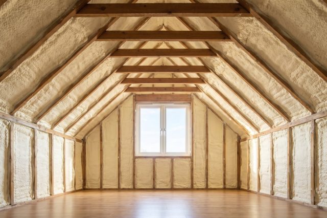 A British loft where thick pale yellow spray foam completely encases the rafters and roof underside, seen in strong soft daylight from a gable window