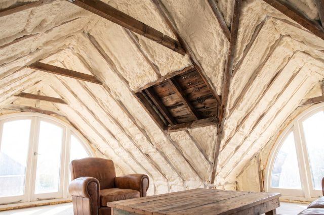 A British loft where dense pale foam completely hides the roof timbers, with one small area cut open showing dark damp timber inside