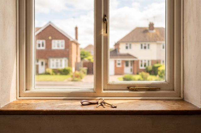 A set of house keys resting on a sunlit windowsill inside a British home, with a softly blurred residential street visible through the glass
