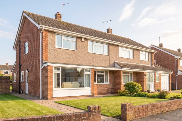 A tidy British 1960s semi-detached house with a shallow tiled roof, large picture window and neat front lawn, in clear bright sunshine