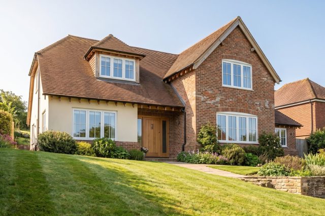A British detached house in warm sunshine seen from the front garden, handsome and well-kept, with a soft downward-sloping foreground lawn