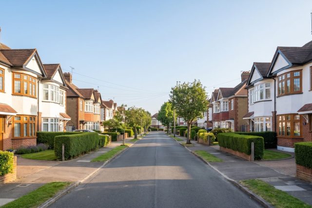 A British suburban street of 1930s bay-fronted semis in bright morning sunshine, clipped hedges, red brick and white render