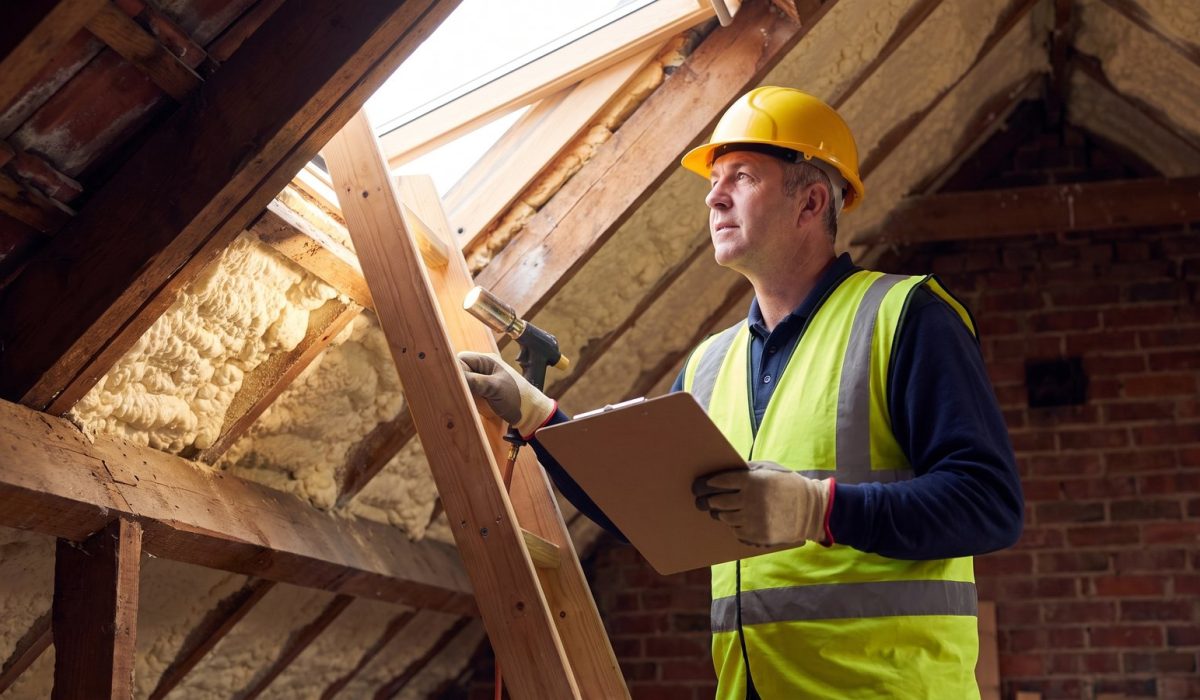 A surveyor in a yellow hard hat and hi-vis vest standing on a loft ladder with head and shoulders up in a sunlit British loft