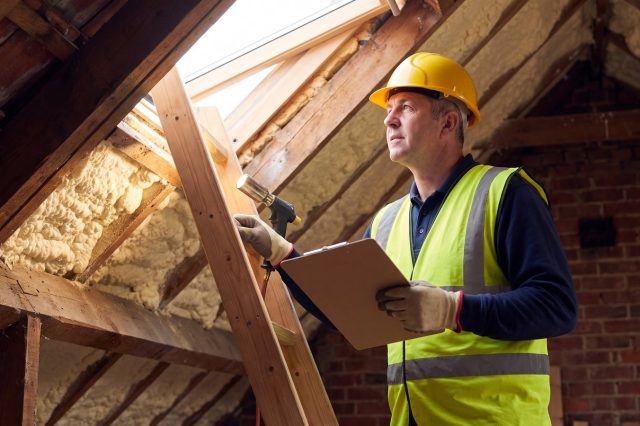 A surveyor in a yellow hard hat and hi-vis vest standing on a loft ladder with head and shoulders up in a sunlit British loft