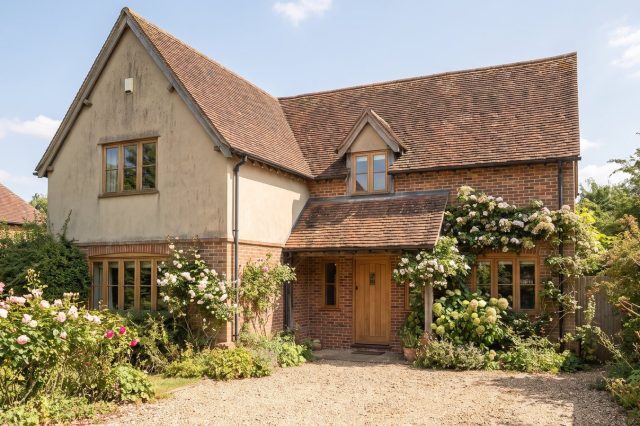 An older British house exterior in soft warm sunshine, slightly dated but well-built, with mature garden planting and a quiet, still atmosphere
