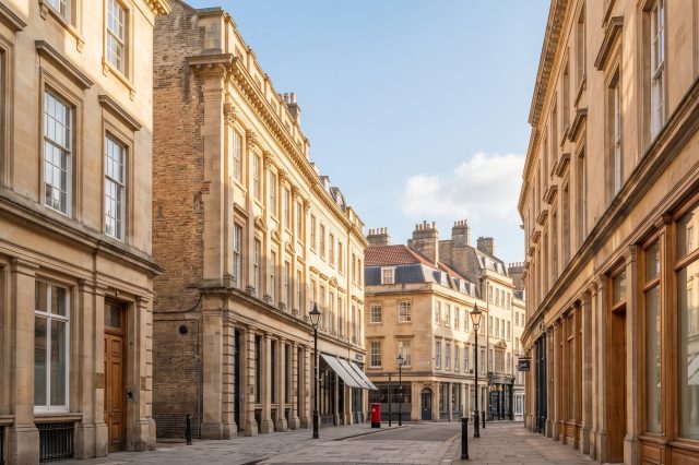 A sunny British city street with solid stone-fronted commercial buildings and tall sash windows, warm daylight raking across the facades