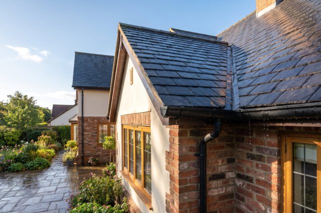 A British house exterior in bright sunshine after light rain, roof tiles glistening, water dripping gently from a gutter, clear sky returning