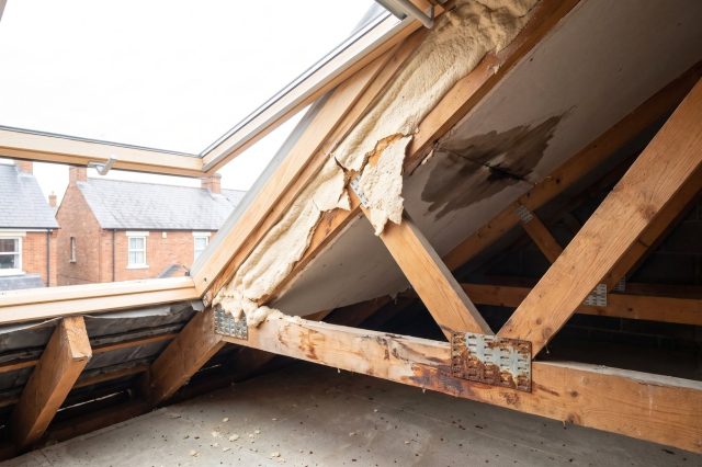 A British loft interior showing several problems at once: pale foam cracking away from a rafter, a dark damp patch, and a rusted nail plate