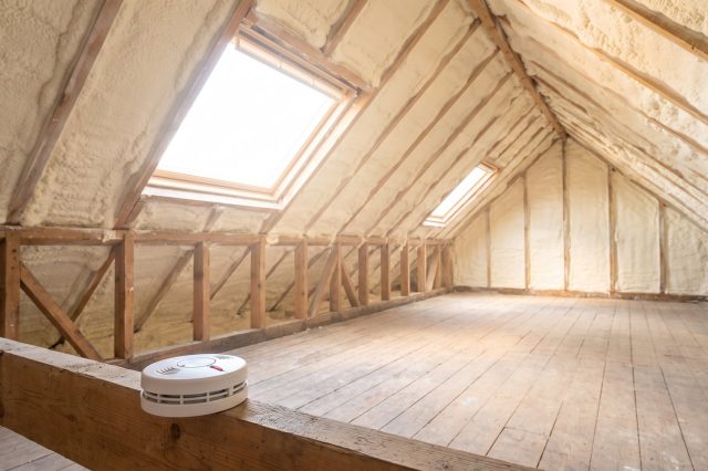 A British loft with pale spray foam on the rafters and a mains-powered smoke alarm mounted on a joist in the foreground