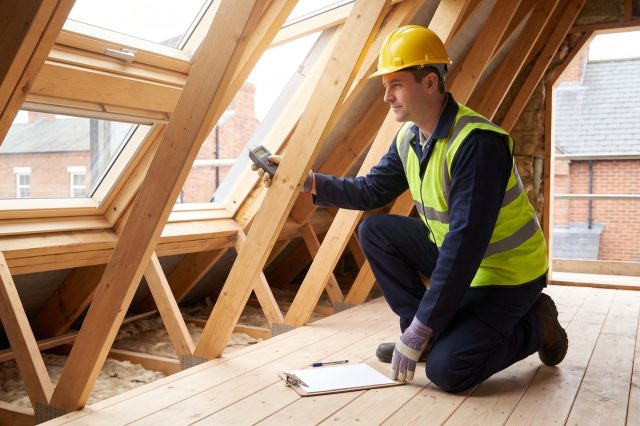 A surveyor in a yellow hard hat and hi-vis vest kneeling on loft boards in a sunlit British loft, using a moisture meter against a rafter