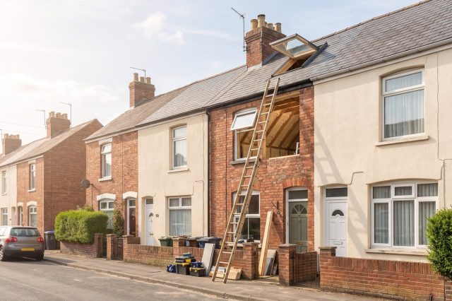 A row of modest British houses in bright sunshine, one with a loft window open and a ladder against the wall, a scene of past retrofit work