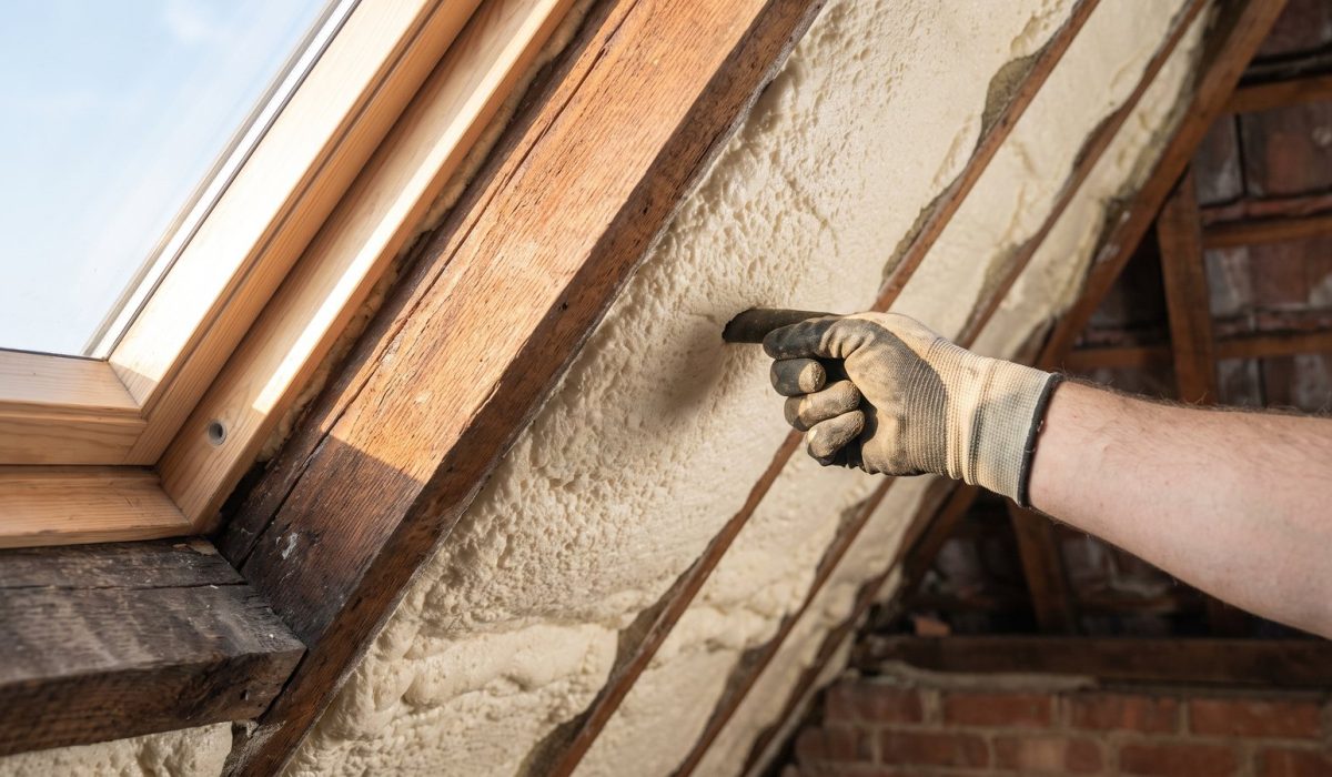 A close-up of a gloved hand pressing a finger into pale foam on a British loft rafter to test its firmness