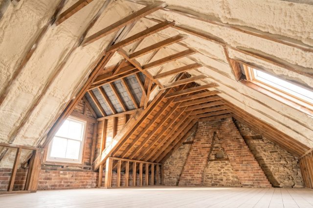 A British loft with pale foam on the rafters seen from a low angle, one clean area already stripped back to bare timber, presenting an open question